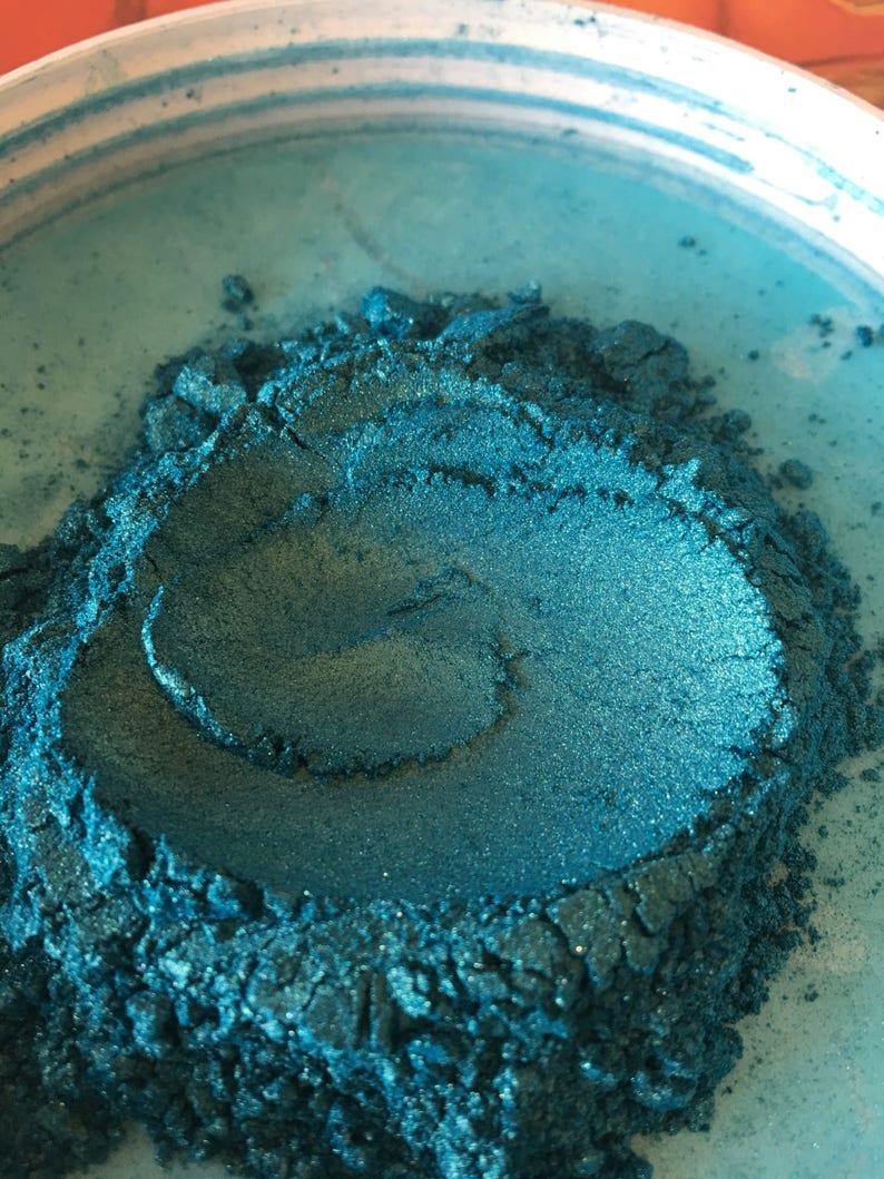 May include: Close-up of a container filled with teal-colored pigment powder. The powder has a shimmering, metallic finish and a textured appearance. The container is white with a teal interior.