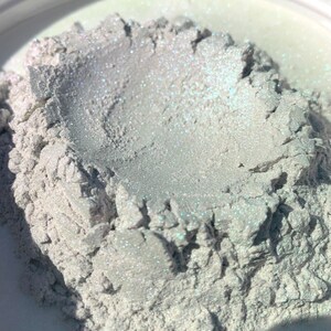 Puede incluir: Primer plano de un mont&oacute;n de polvo nacarado con un efecto brillante e iridiscente. El polvo es de un color claro y neutro con sutiles toques de azul y verde. La textura parece granular y ligeramente &aacute;spera, lo que sugiere un material cosm&eacute;tico o artesanal.