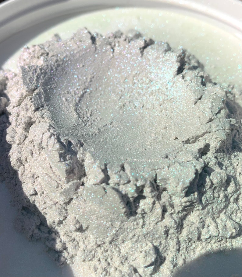 Puede incluir: Primer plano de un mont&oacute;n de polvo nacarado con un efecto brillante e iridiscente. El polvo es de un color claro y neutro con sutiles toques de azul y verde. La textura parece granular y ligeramente &aacute;spera, lo que sugiere un material cosm&eacute;tico o artesanal.