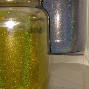 May include: Two clear glass jars filled with glitter. One jar contains gold glitter, and the other contains silver glitter. Both jars have white lids. The jars are displayed on a clear, tiered shelf.