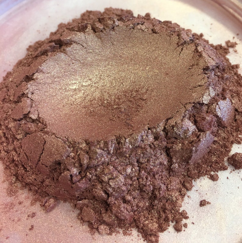May include: A close-up of a pile of shimmering, finely milled powder. The powder has a metallic, bronzy-rose color with lighter, golden highlights. The texture appears soft and loose, suggesting a cosmetic product like eyeshadow or pigment.