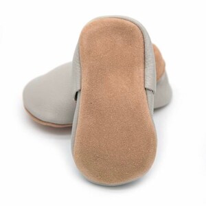 May include: A pair of baby shoes in a light gray color. The soles are a tan suede material. The shoes are made of soft leather and are designed for infants. The shoes are shown on a white background.