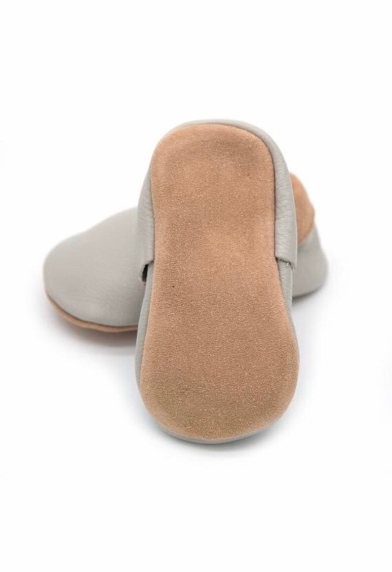 May include: A pair of baby shoes in a light gray color. The soles are a tan suede material. The shoes are made of soft leather and are designed for infants. The shoes are shown on a white background.