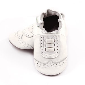 May include: A pair of white leather baby shoes with a classic brogue design. The shoes feature decorative perforations and a three-strap detail on the front. The soft-soled shoes are designed for infants.