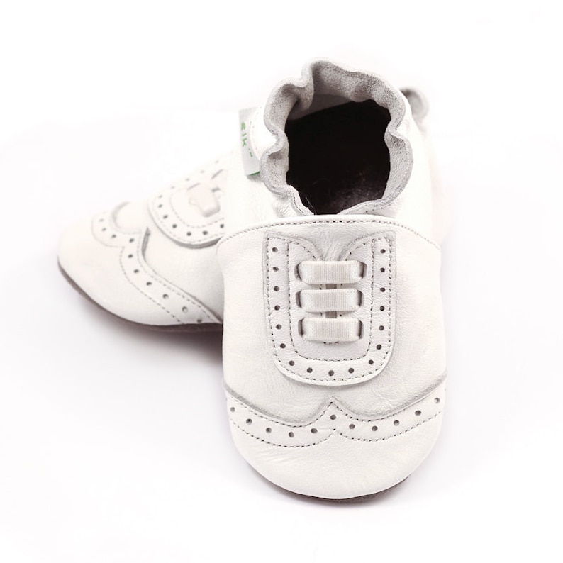 May include: A pair of white leather baby shoes with a classic brogue design. The shoes feature decorative perforations and a three-strap detail on the front. The soft-soled shoes are designed for infants.