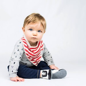 May include: A baby wearing a gray long-sleeved shirt with a small tree pattern, dark blue jeans, and white and black shoes. The baby is also wearing a red and white striped bandana bib.