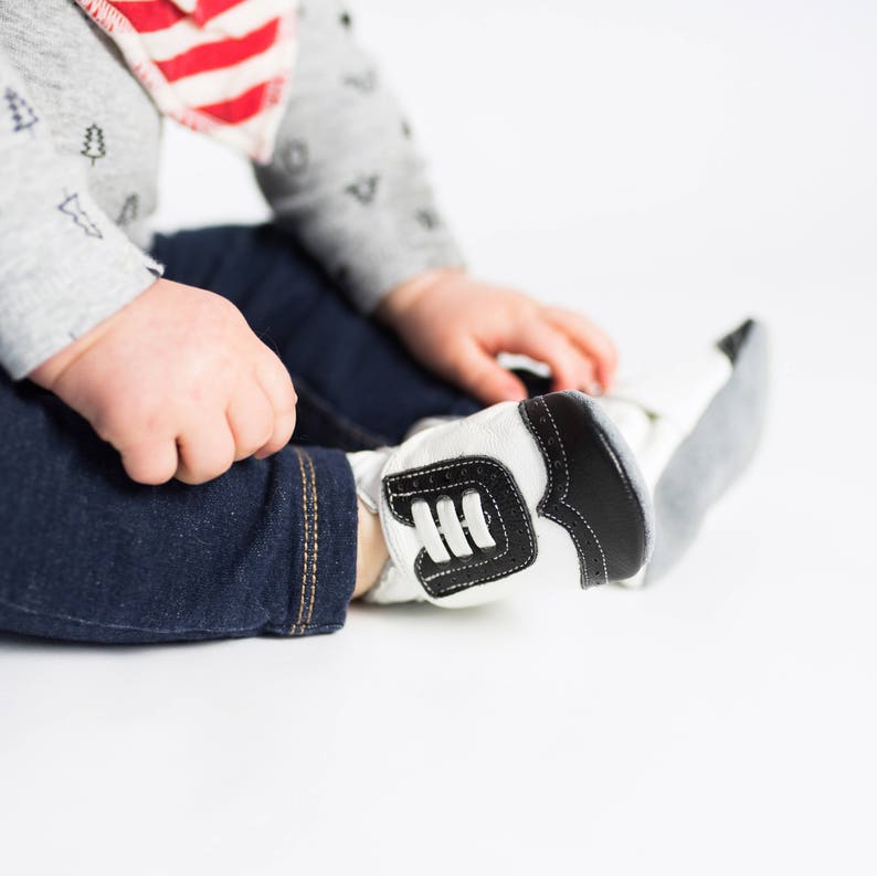 May include: White and black baby shoes with a sneaker design. The shoes have a white base with black accents and a lace-up detail. The baby is wearing dark blue jeans and a gray shirt.