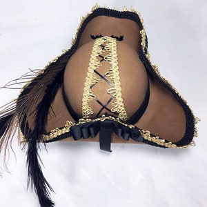 May include: A brown felt pirate hat with black trim and a gold braid design. The hat has a black feather attached to the side.