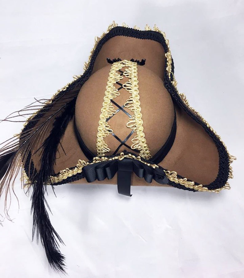 May include: A brown felt pirate hat with black trim and a gold braid design. The hat has a black feather attached to the side.