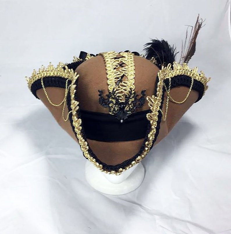 May include: A brown felt hat with gold and black trim. The hat has a black band with a gold and black design. The hat is decorated with a black feather.