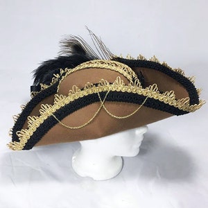 May include: A brown felt pirate hat with black and gold trim and a black feather. The hat has a chain detail and is angled to the side.