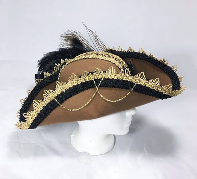 May include: A brown felt pirate hat with black and gold trim and a black feather. The hat has a chain detail and is angled to the side.