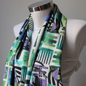 Könnte beinhalten: Ein weißer Schal mit einem geometrischen Muster in Grün-, Blau- und Lilatönen. Das Muster besteht aus Streifen, Quadraten und Dreiecken.
