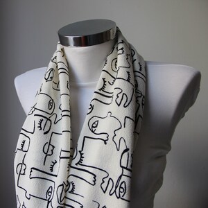 May include: A white scarf with a black and white abstract pattern of faces. The scarf is folded and draped over a white mannequin.