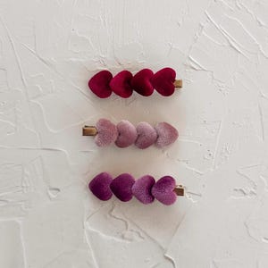 May include: Three velvet heart-shaped hair clips in shades of red, pink, and purple. Each clip features a row of three hearts attached to a gold-colored metal barrette. The clips are arranged on a textured white surface.