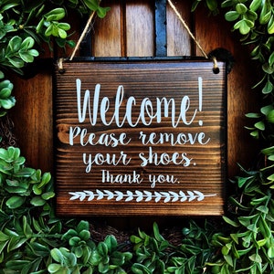 May include: A wooden sign with the text "Welcome! Please remove your shoes. Thank you." in white lettering. The sign is hanging from a string and is surrounded by green foliage.
