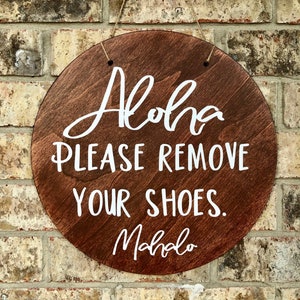 May include: A round wooden sign with the text "Aloha Please Remove Your Shoes. Mahalo" written in white paint.
