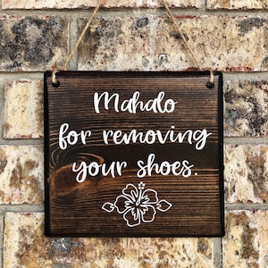 May include: A dark brown wooden sign with white text that reads "Mahalo for removing your shoes." A white hibiscus flower is below the text.
