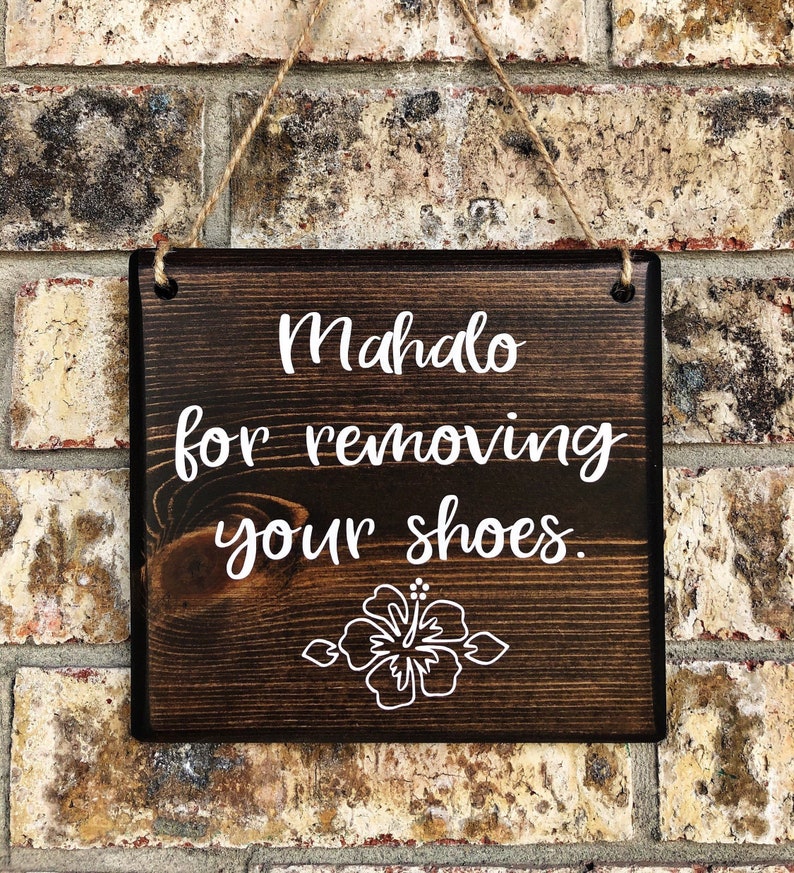 May include: A dark brown wooden sign with white text that reads "Mahalo for removing your shoes." A white hibiscus flower is below the text.