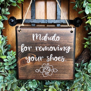 May include: A dark brown wooden sign with white text that reads "Mahalo for removing your shoes." A white hibiscus flower is centered below the text. The sign is hanging from a wreath of green leaves.