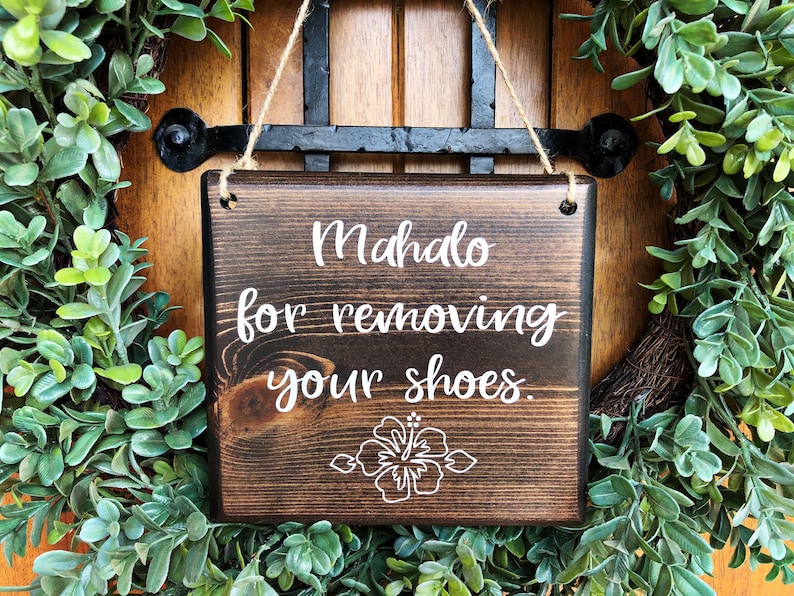 May include: A dark brown wooden sign with white text that reads "Mahalo for removing your shoes." A white hibiscus flower is centered below the text. The sign is hanging from a wreath of green leaves.