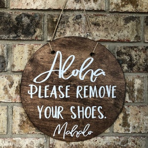 May include: A round wooden sign with the text "Aloha Please Remove Your Shoes. Mahalo" in white lettering.