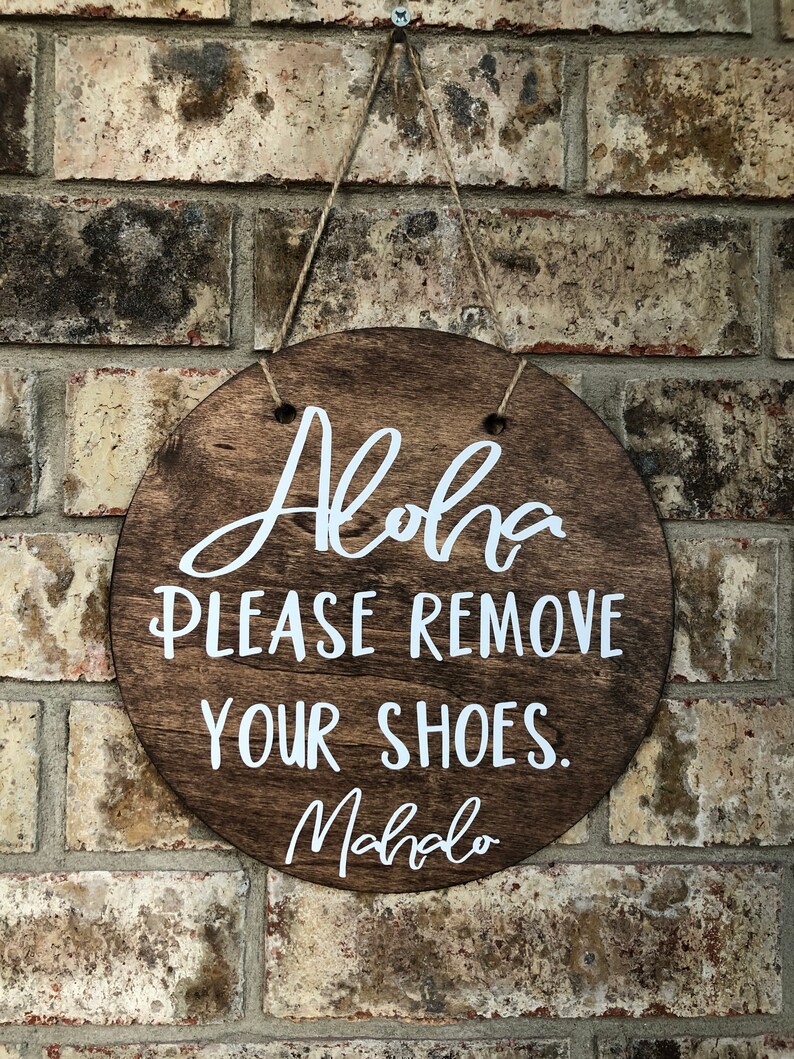 May include: A round wooden sign with the text "Aloha Please Remove Your Shoes. Mahalo" in white lettering.