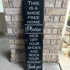 May include: Black wooden sign with white text that reads "This is a shoe free home. Please kick off your shoes and rock your socks! Thank you!"