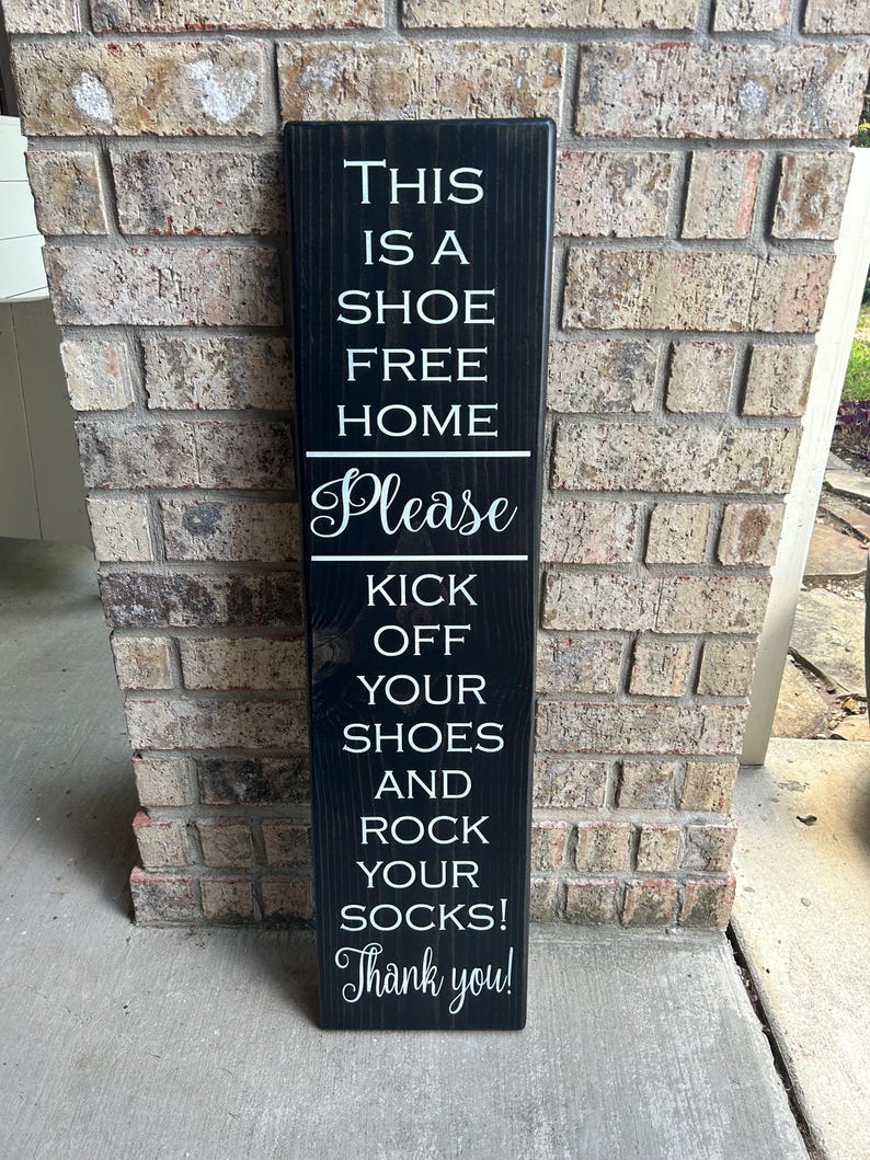 May include: Black wooden sign with white text that reads "This is a shoe free home. Please kick off your shoes and rock your socks! Thank you!"