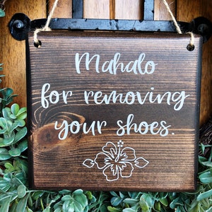 May include: A dark brown wooden sign with white text that reads "Mahalo for removing your shoes." A white hibiscus flower is drawn at the bottom of the sign.