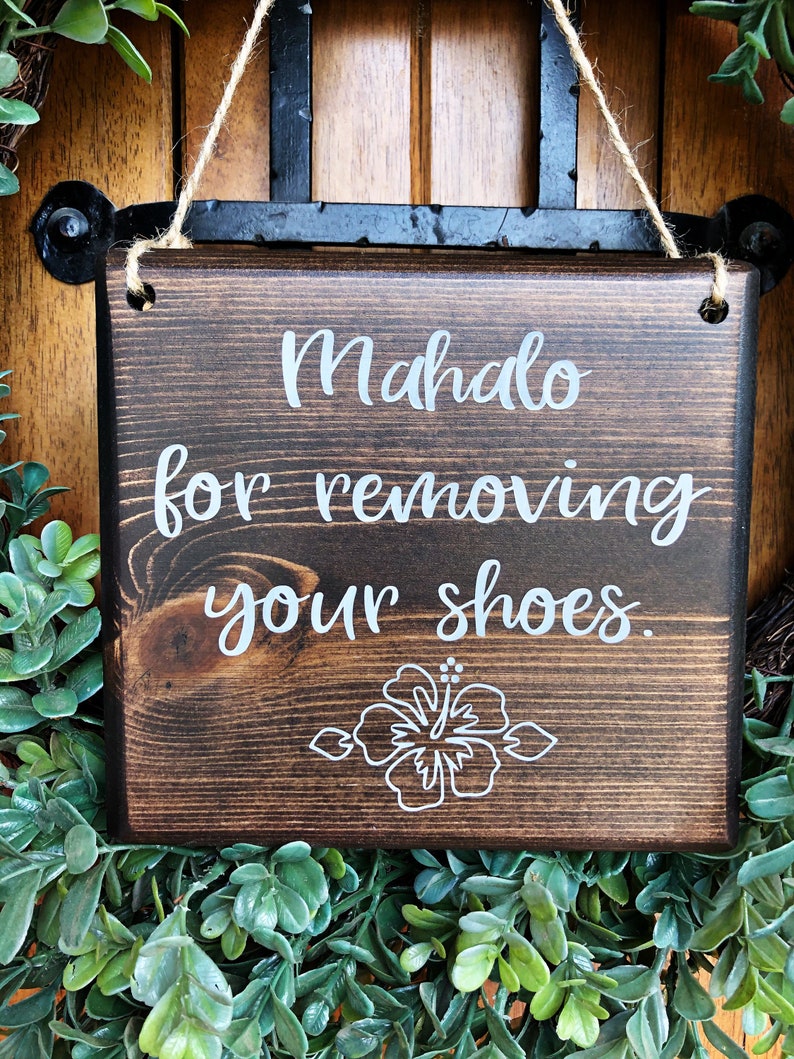 May include: A dark brown wooden sign with white text that reads "Mahalo for removing your shoes." A white hibiscus flower is drawn at the bottom of the sign.