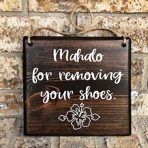 May include: A dark brown wooden sign with white text that reads "Mahalo for removing your shoes." A white hibiscus flower is below the text.
