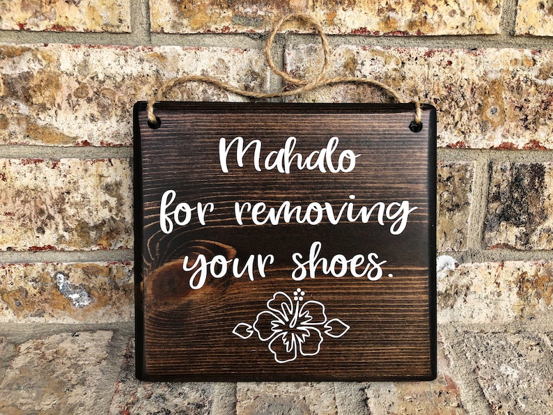 May include: A dark brown wooden sign with white text that reads "Mahalo for removing your shoes." A white hibiscus flower is below the text.