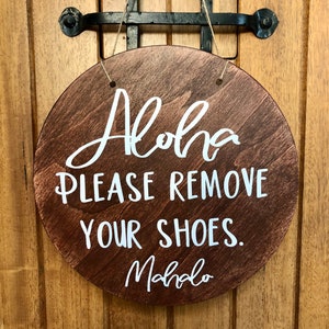 May include: A round wooden sign with the text "Aloha Please Remove Your Shoes. Mahalo" in white lettering.