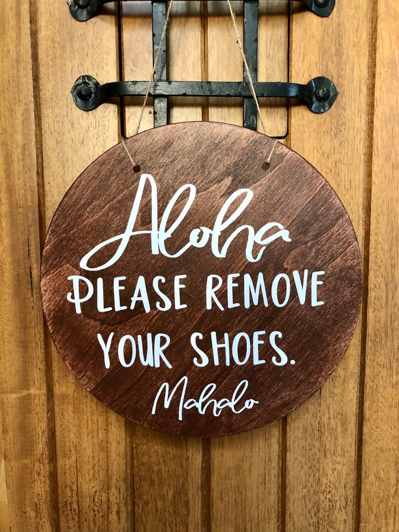 May include: A round wooden sign with the text "Aloha Please Remove Your Shoes. Mahalo" in white lettering.