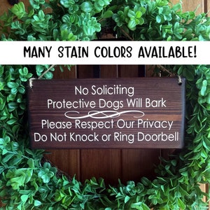 May include: A wooden sign with the text "No Soliciting, Protective Dogs Will Bark, Please Respect Our Privacy, Do Not Knock or Ring Doorbell". The sign is brown and has white text. The sign is hanging on a door with a green wreath.