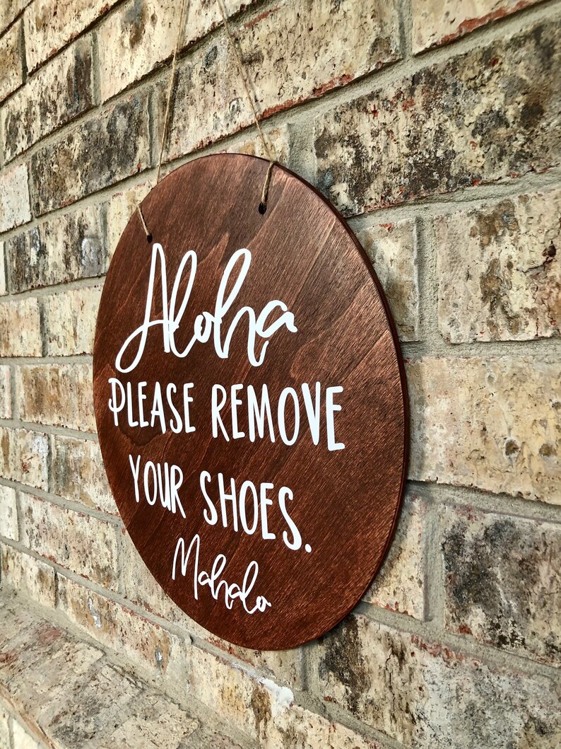 May include: A round wooden sign with the text "Aloha Please Remove Your Shoes. Mahalo" written in white paint.