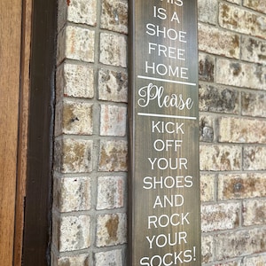 May include: A brown wooden sign with white text that reads "This is a shoe free home. Please kick off your shoes and rock your socks! Thank you!"