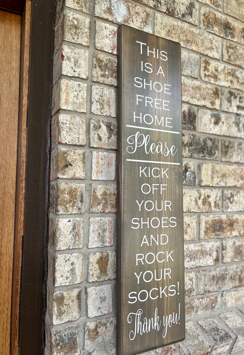May include: A brown wooden sign with white text that reads "This is a shoe free home. Please kick off your shoes and rock your socks! Thank you!"