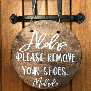 May include: A round wooden sign with the text "Aloha Please Remove Your Shoes. Mahalo" in white lettering.
