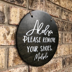 May include: A round wooden sign with the text "Aloha Please Remove Your Shoes. Mahalo" in white lettering on a black background.