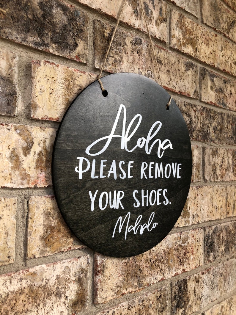May include: A round wooden sign with the text "Aloha Please Remove Your Shoes. Mahalo" in white lettering on a black background.