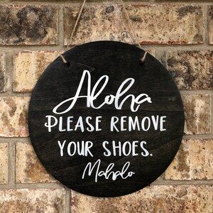 May include: A round wooden sign with the text "Aloha Please Remove Your Shoes. Mahalo" written in white paint.