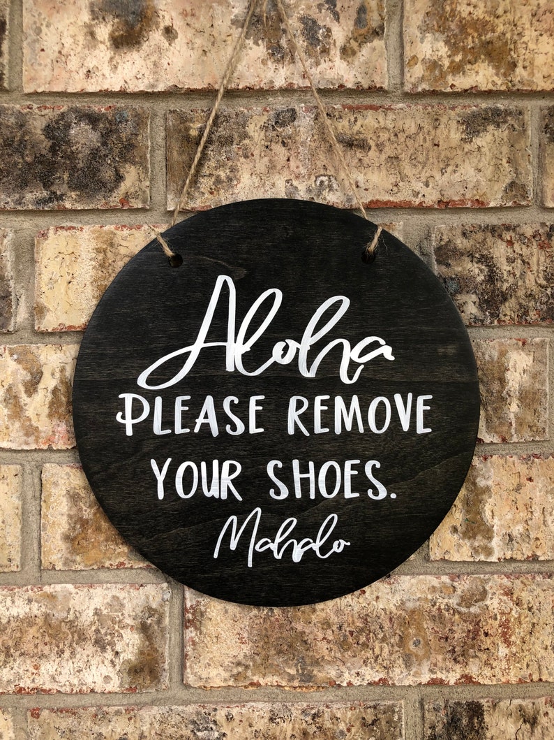 May include: A round wooden sign with the text "Aloha Please Remove Your Shoes. Mahalo" written in white paint.