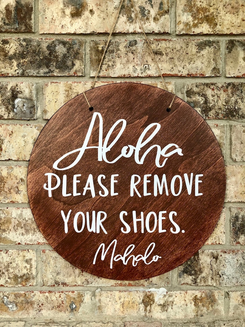 May include: A round wooden sign with the text "Aloha Please Remove Your Shoes. Mahalo" written in white paint.