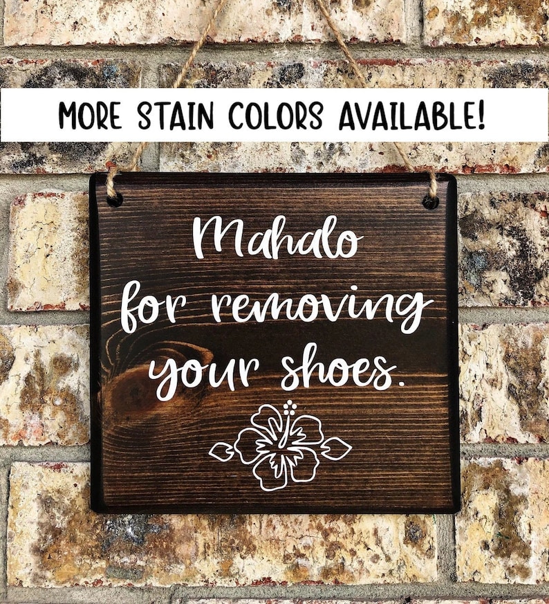 May include: A dark brown wooden sign with white text that reads "Mahalo for removing your shoes." A white hibiscus flower is below the text. The sign has two holes at the top for hanging.