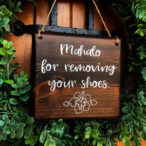 May include: A wooden sign with the text "Mahalo for removing your shoes." in white lettering. The sign has a brown stain and a white hibiscus flower design.