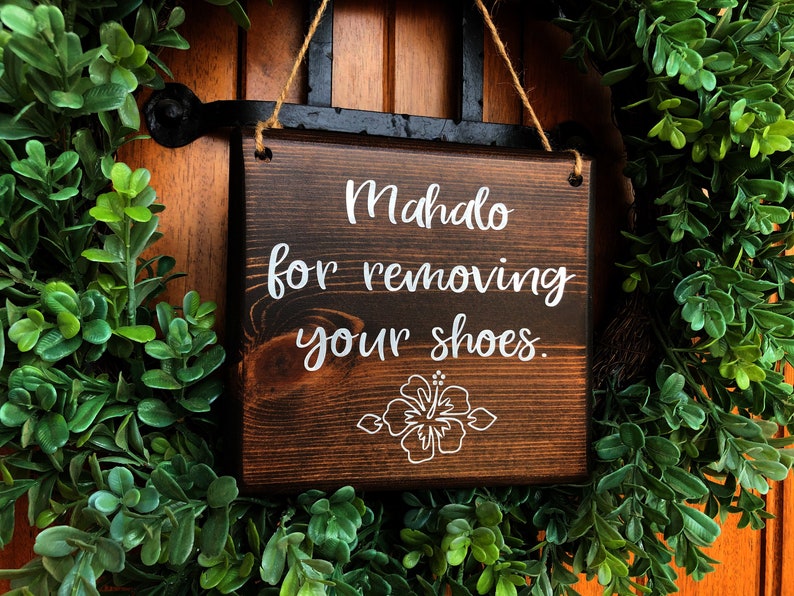 May include: A wooden sign with the text "Mahalo for removing your shoes." in white lettering. The sign has a brown stain and a white hibiscus flower design.