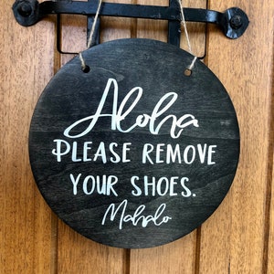 May include: A round wooden sign with a black background and white text that reads "Aloha Please Remove Your Shoes. Mahalo". The sign is hanging from a string.