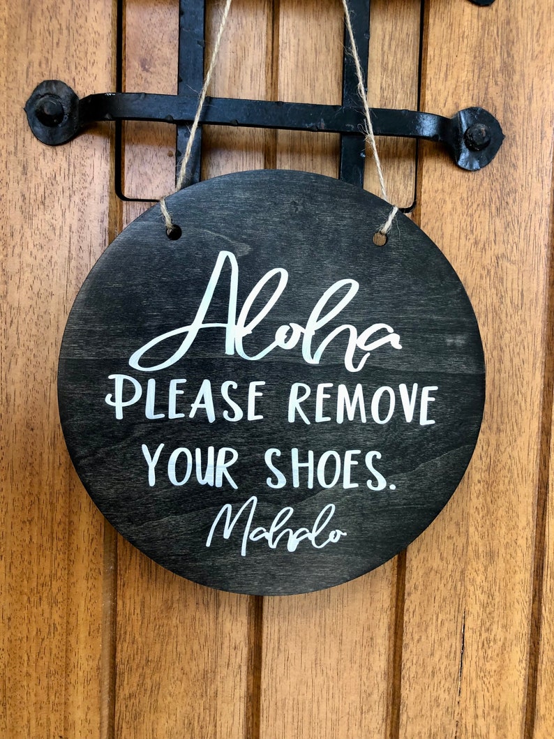 May include: A round wooden sign with a black background and white text that reads "Aloha Please Remove Your Shoes. Mahalo". The sign is hanging from a string.